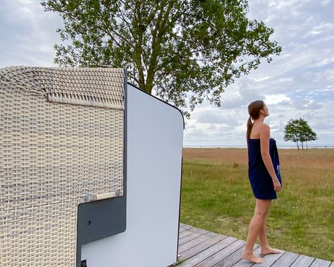 Eine nur mit Handtuch bekleidete Frau steht auf einem Holzpodest vor einer Strandkorbsauna und blickt in die Natur