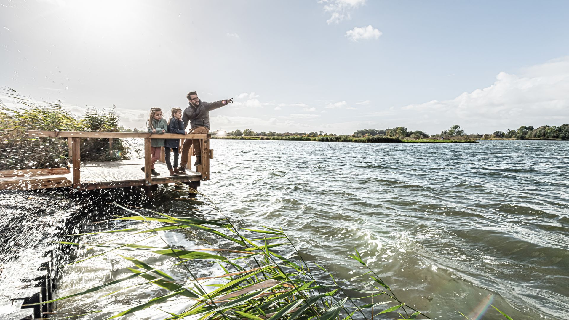 Kleiner Steg an einem See auf dem ein Vater mit zwei Kindern steht. Der Vater zeigt in die Ferne