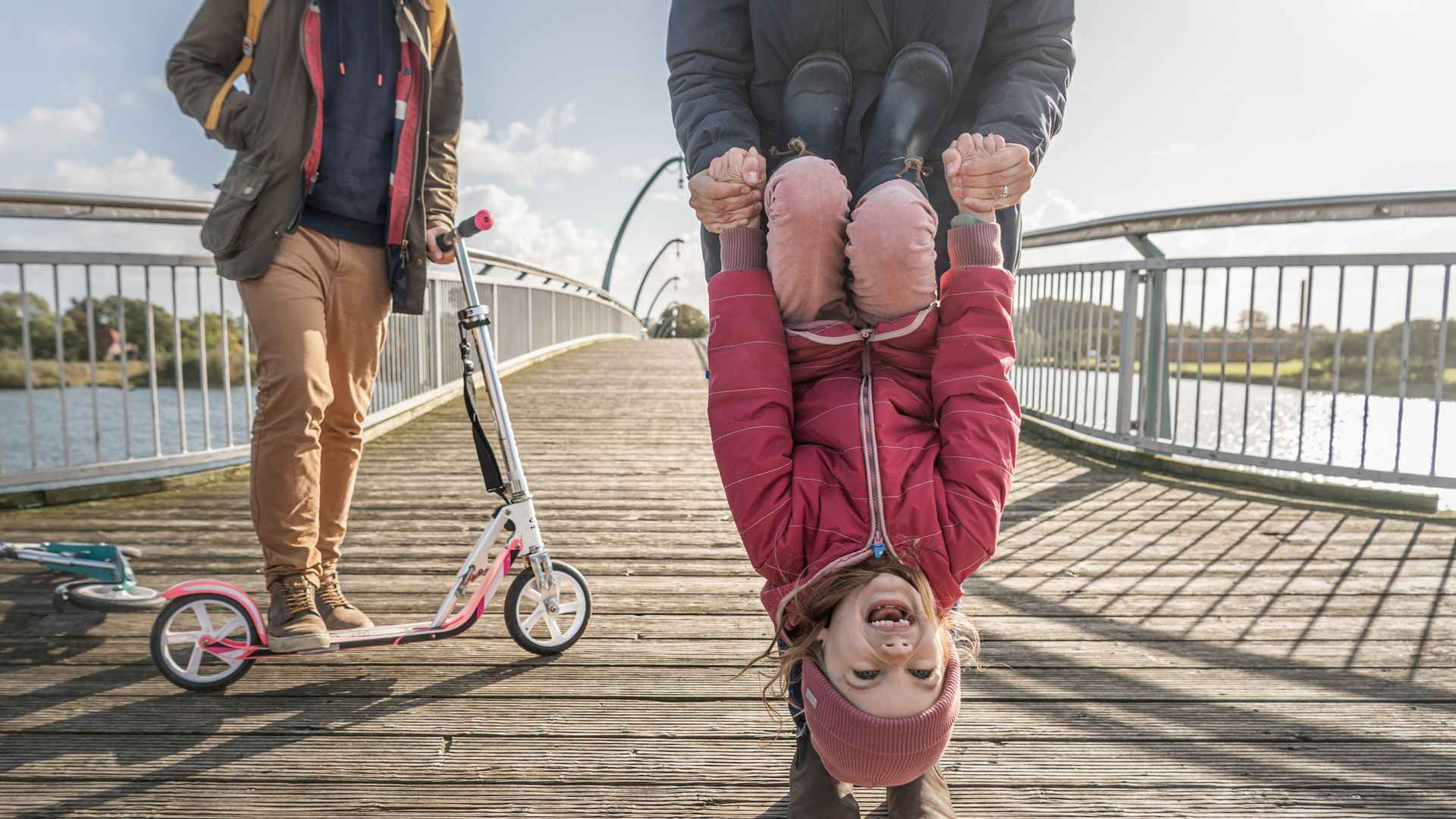 Ein Kind turnt auf einer Holzbrücke an seiner Mutter. Im Hintergrund steht ein Mann mit einem Tretroller