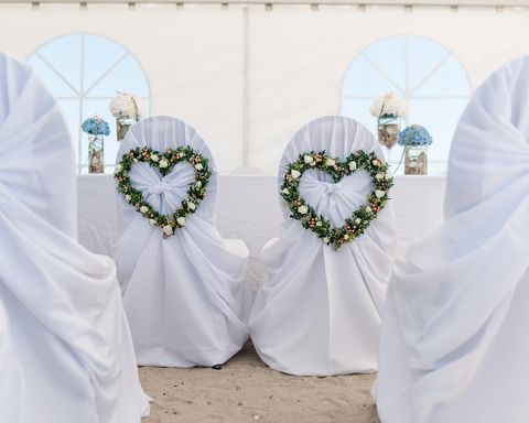 Foto einer Hochzeitslocation. Stühle mit weißen Überwurf und mit Herzen aus Blumen und Zweigen an der Rückseite. 