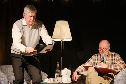 Ein Mann sitzt mit einem Buch in einem Sessel, ein anderer setzt sich gerade neben ihn auf einen Sessel