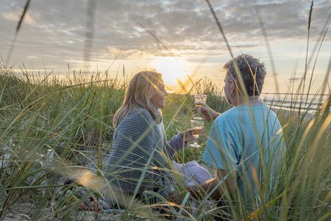 Ein Pärchen sitzt bei Sonnenuntergang in den Dünen und stößt mit Sekt an
