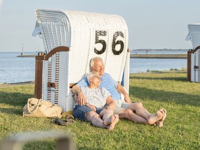 Ein älteres Pärchen sitzt mit dem Rücken an einen Strandkorb gelehnt auf einer Wiese 