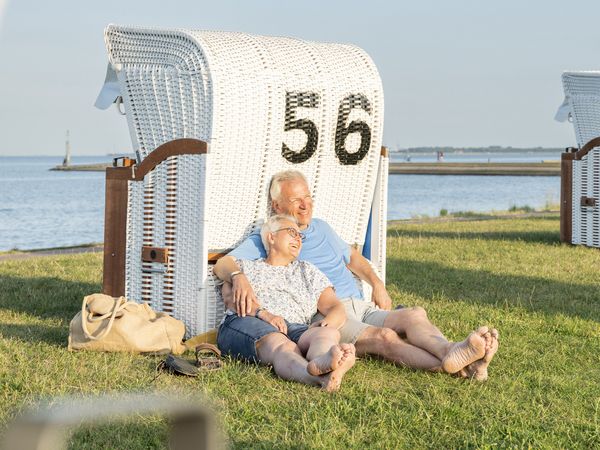 Ein älteres Pärchen sitzt mit dem Rücken an einen Strandkorb gelehnt auf einer Wiese 