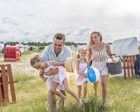 Eine Familie mit zwei Kindern läuft über eine Wiese mit Strandkörben. Der Vater albert mit einem der Kinder rum und hebt es hoch